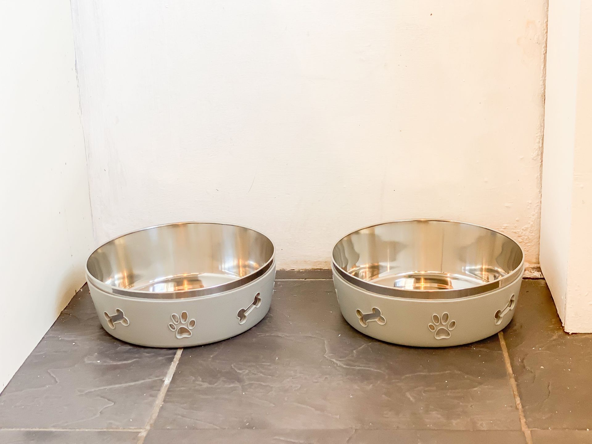 Two stainless steel pet bowls, one gray and one gold, on a tiled floor by a wall