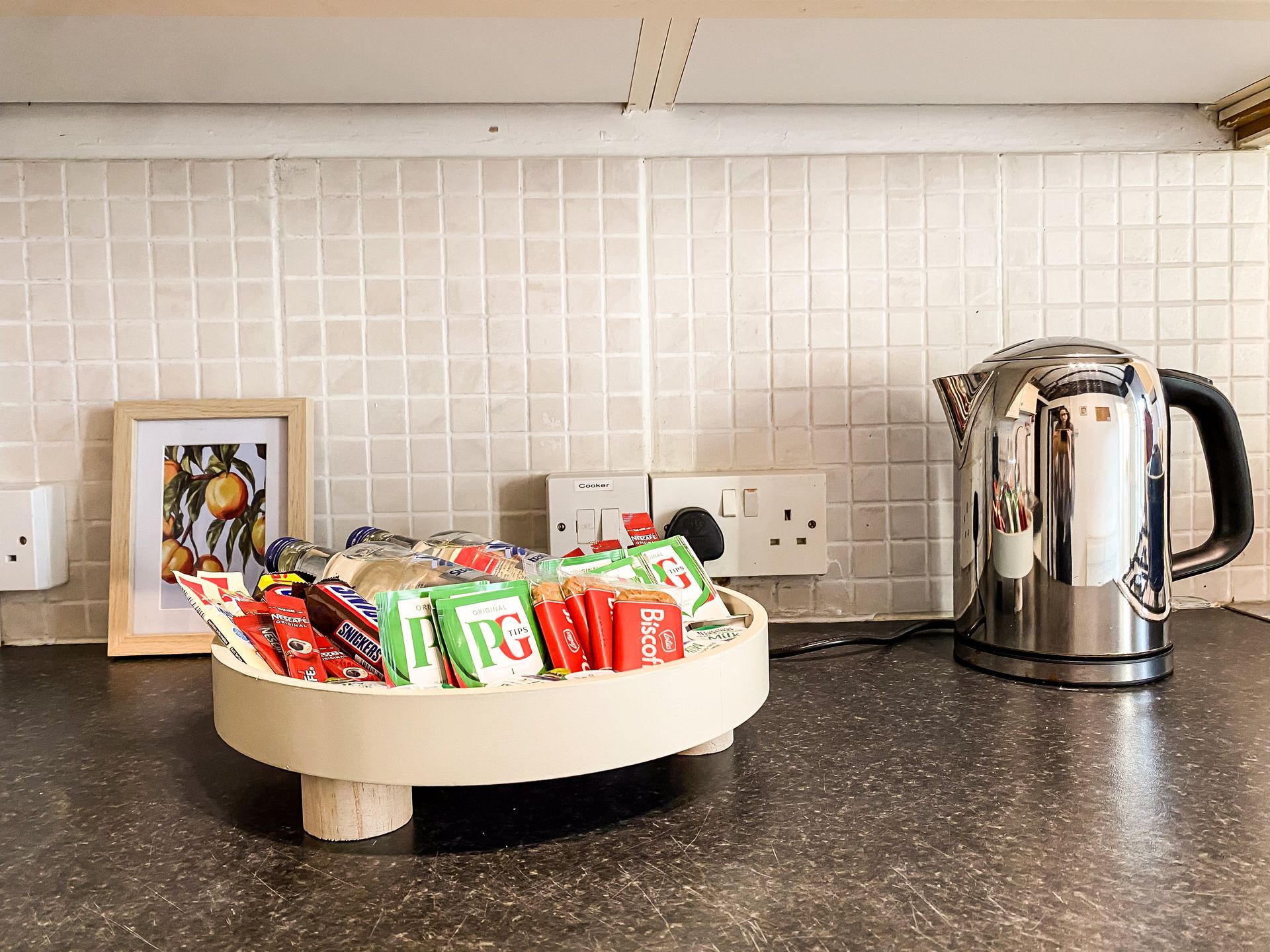 Kitchen counter with a tray of snack bars, a kettle, and a framed photo against a tiled backsplash
