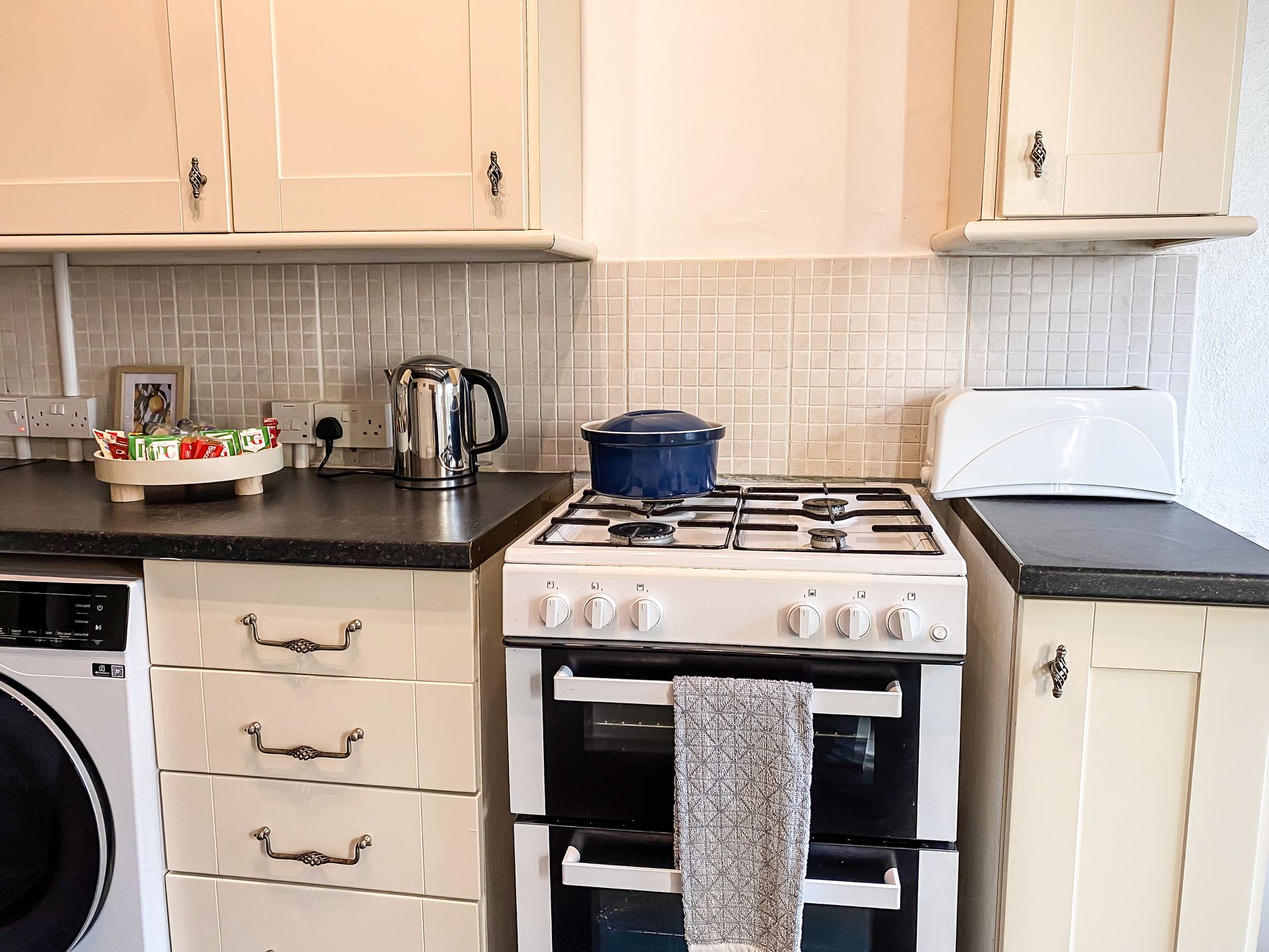 Small kitchen with white cabinets, black countertops, stove, kettle, and washing machine; towel hangs on oven handle.
