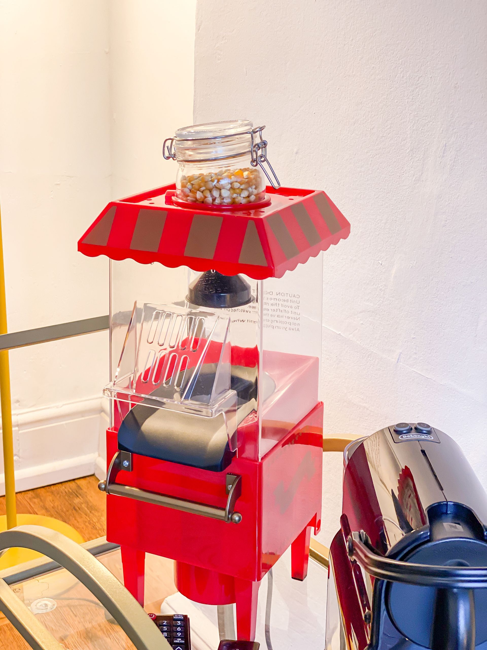 Red popcorn machine on a floor beside a silver suitcase, with a striped top and clear bin.