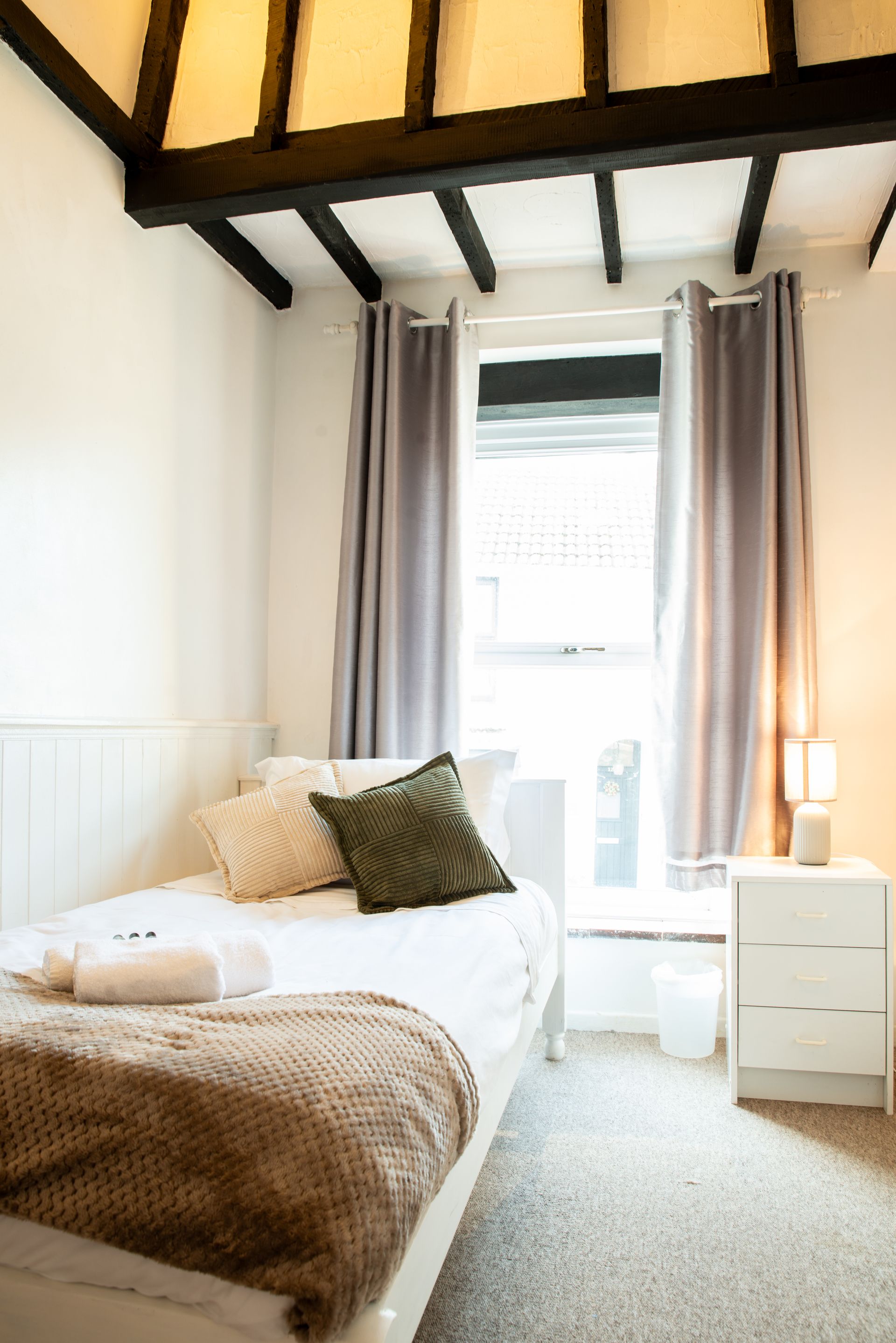Bright bedroom with a single bed, cream curtains, and a nightstand beside a window.