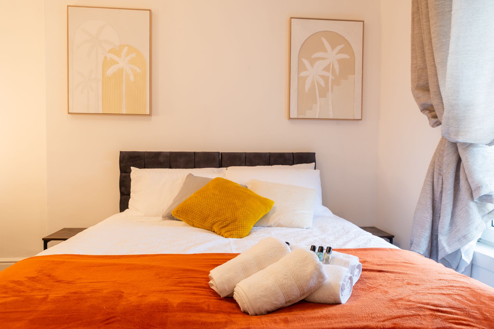 Cozy bedroom with white bedding, orange throw blanket, yellow pillow, and framed palm prints on the wall