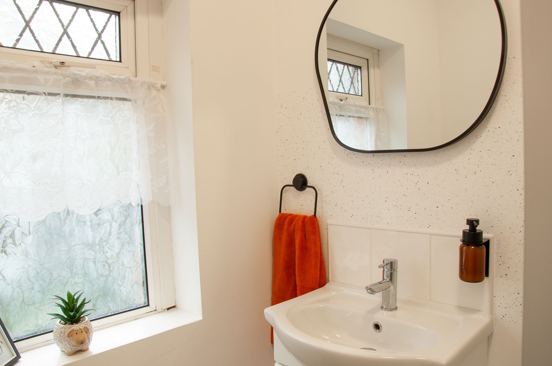 Bright bathroom vanity with white sink, round mirror, orange towel, soap dispenser, and frosted window.