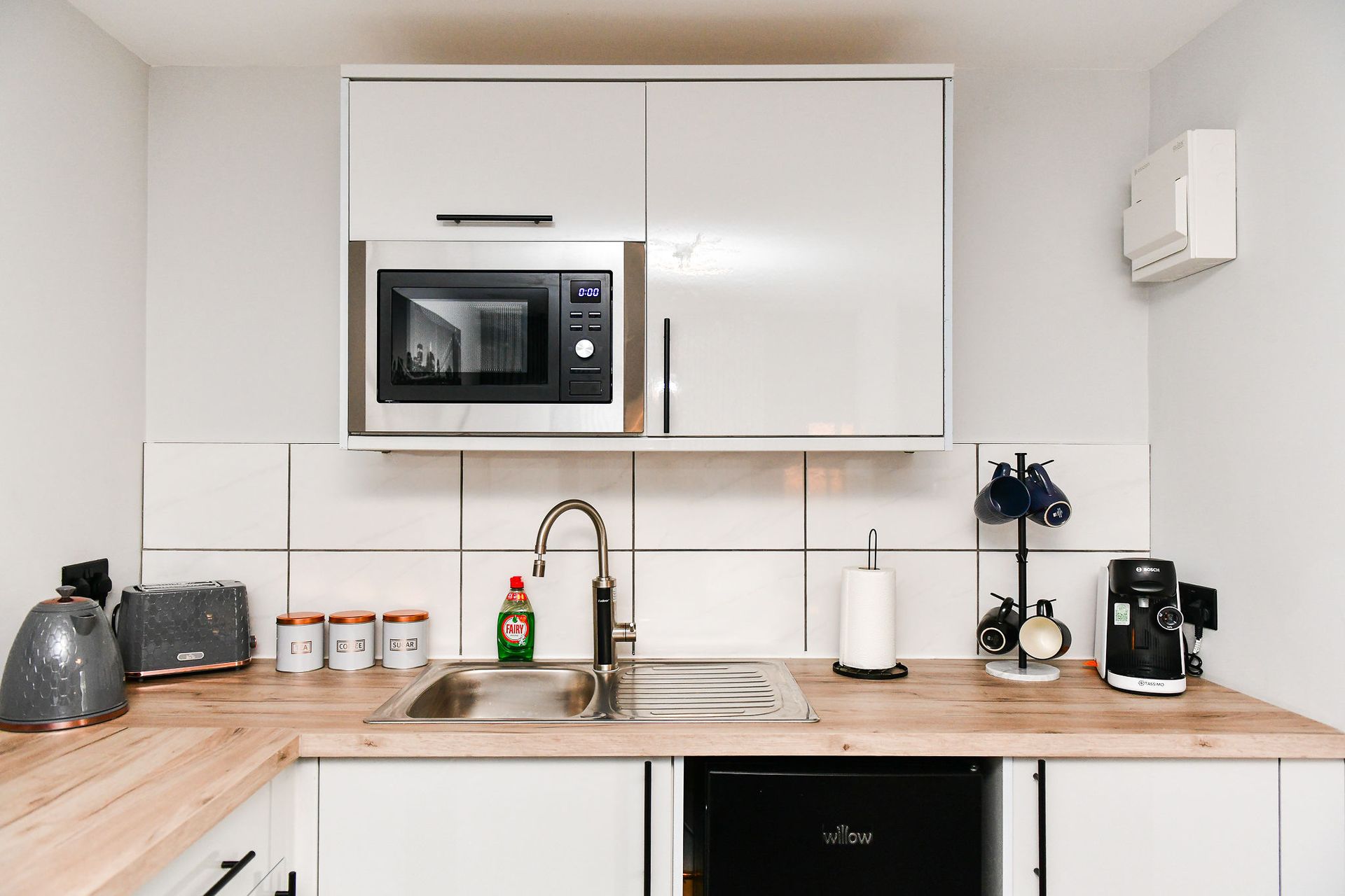 Modern white kitchen with wooden countertop, microwave, sink, kettle, coffee maker, and small decor items