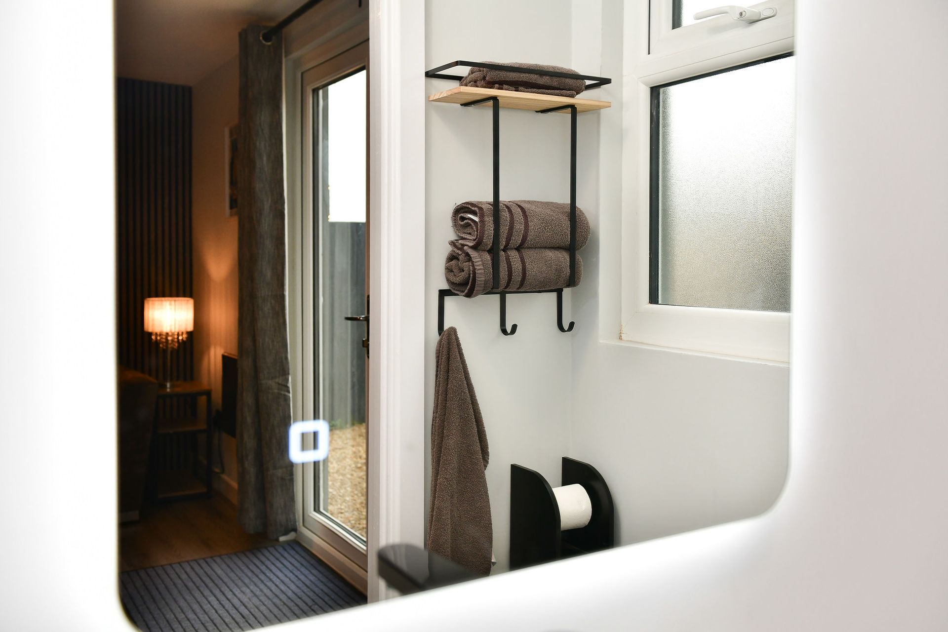 Modern bathroom with towel rack, frosted window, mirror, and doorway to a lit bedroom
