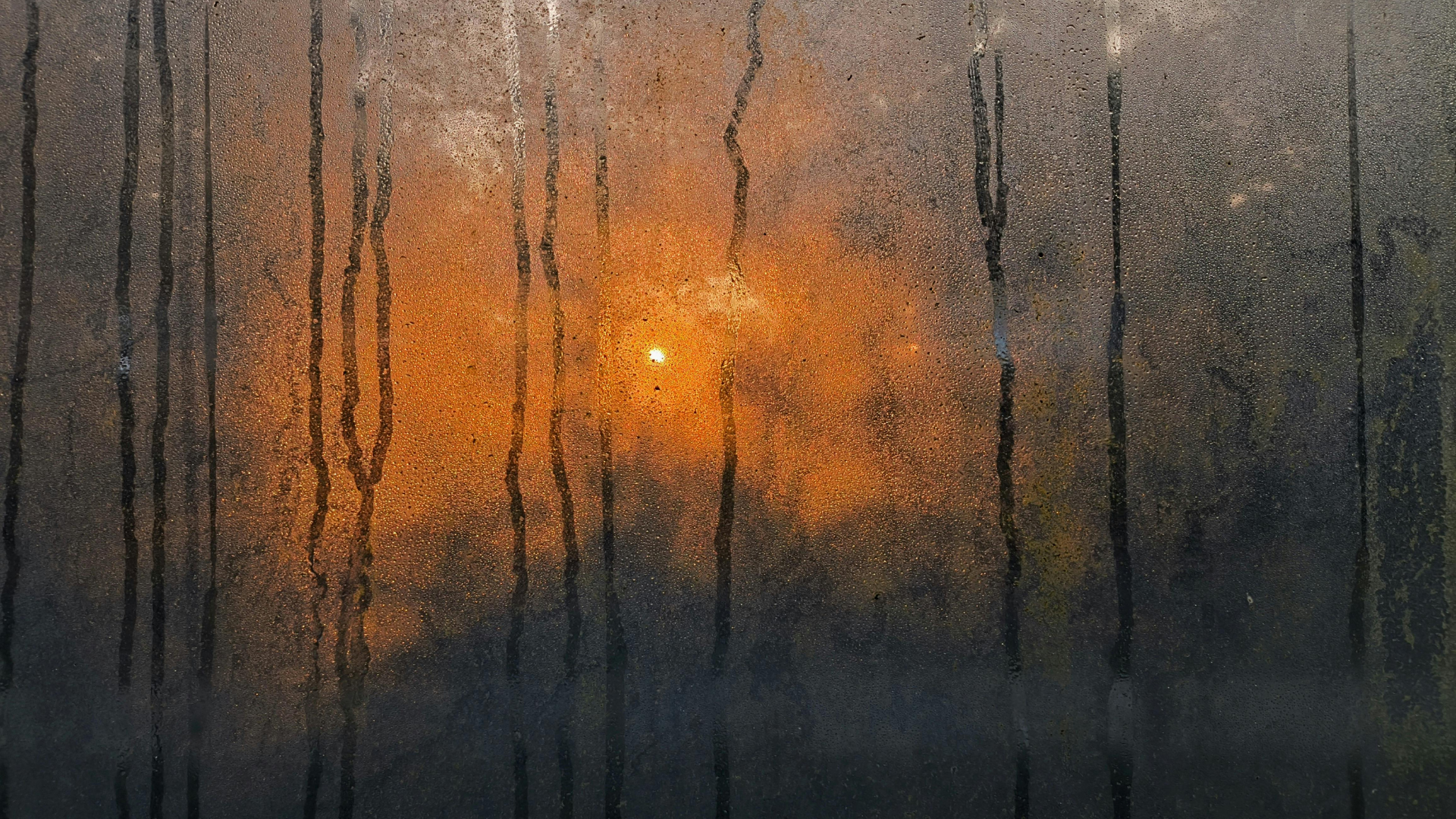Sunset through a condensation-covered window; sun glows orange, vertical water streaks dark against the light.