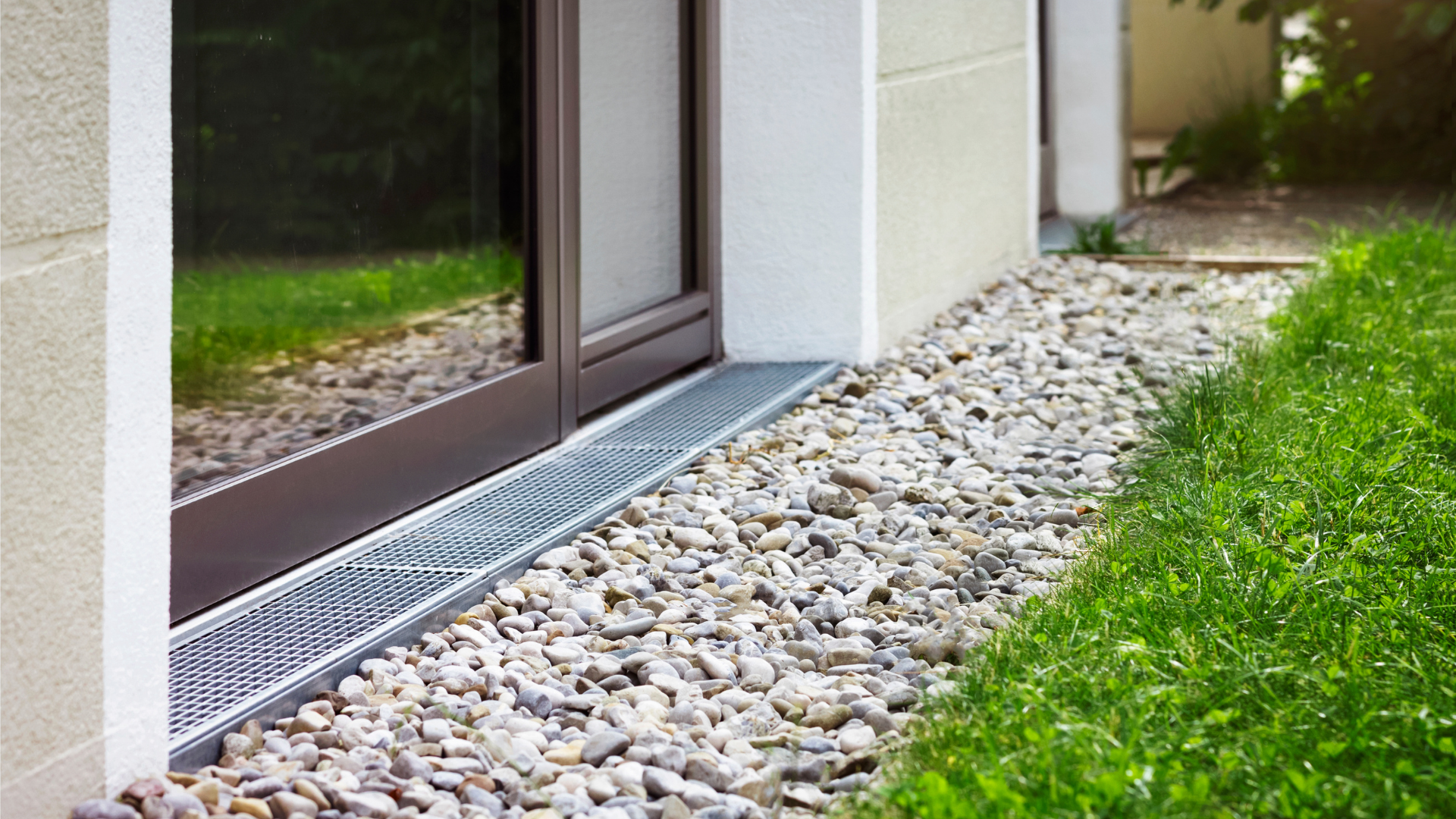 Gravel drainage channel along the base of a building's window, next to a patch of green grass.
