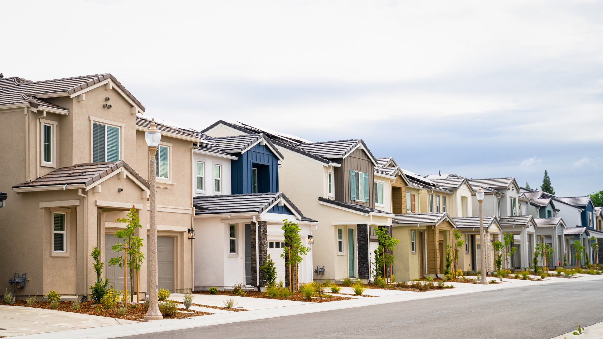 A row of modern, multi-story suburban houses in neutral tones lining a paved street under an overcast sky.