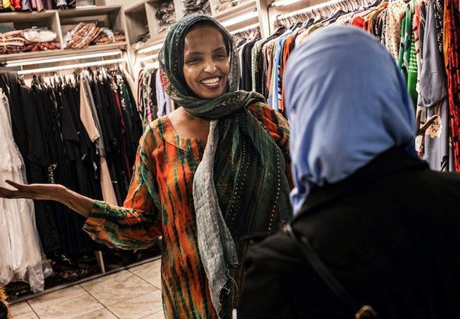 Woman in colorful dress smiles, talking to another woman in a blue hijab inside a clothing store.