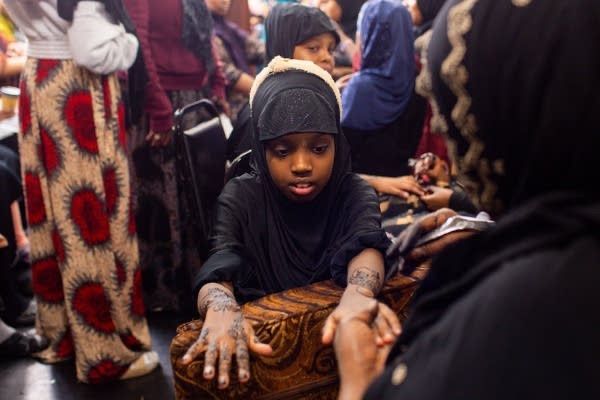Young girl with henna, wearing black hijab, is interacting with an older woman in a crowded setting.