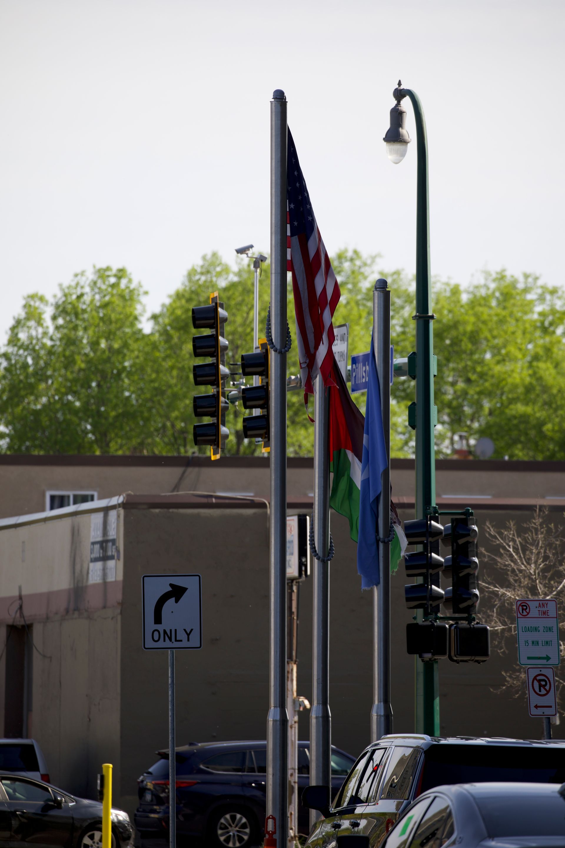 Flags on poles: US flag, green, blue. Street with traffic lights, cars, building, trees.