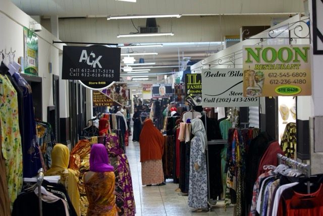 Indoor market with clothing racks and shoppers, various store signs.