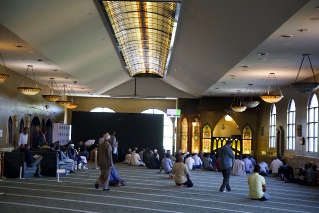People praying inside a mosque with stained glass windows, a large skylight, and green carpet.