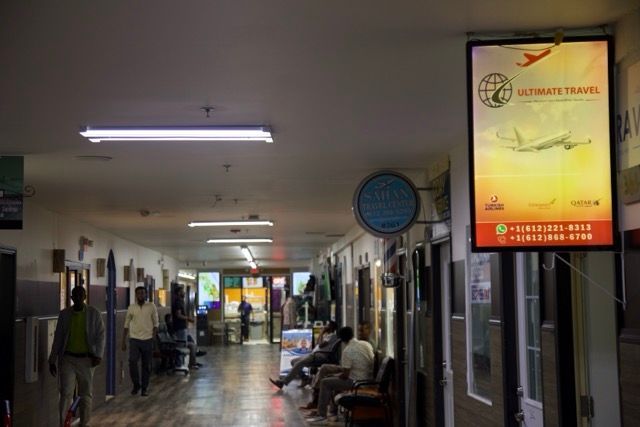 Hallway with travel agency advertisement; people sitting, walking; fluorescent lighting.