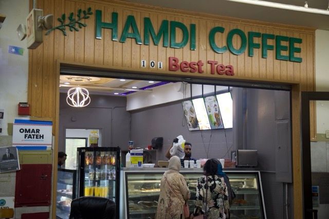 Hamdi Coffee shop with patrons, offering coffee and tea. Green sign above a doorway, interior visible.