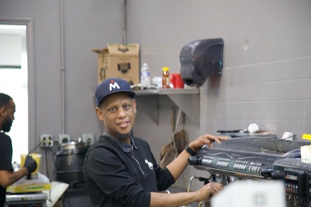 Man in black shirt and hat at coffee machine. Another person in background.