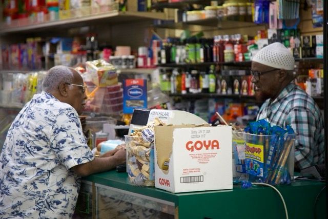 Two men in a convenience store, one at the counter buying items. Shelves with products in the background.