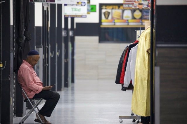 Man in pink shirt sits on a chair, looking at his phone. Clothes hang on a rack in a hallway.