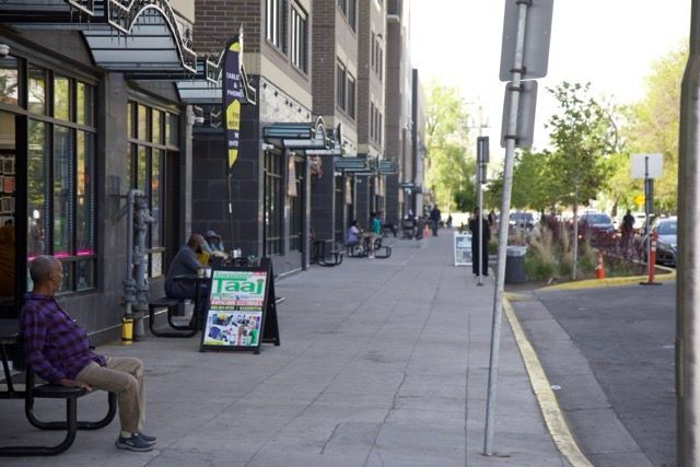 Sidewalk cafe: People seated outdoors, eating and drinking. Buildings in the background. Sunny day.