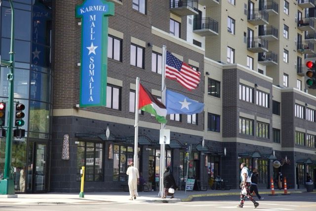 Exterior of Karmel Mall in Minneapolis, with US, Palestinian, and Somali flags. People cross the street.