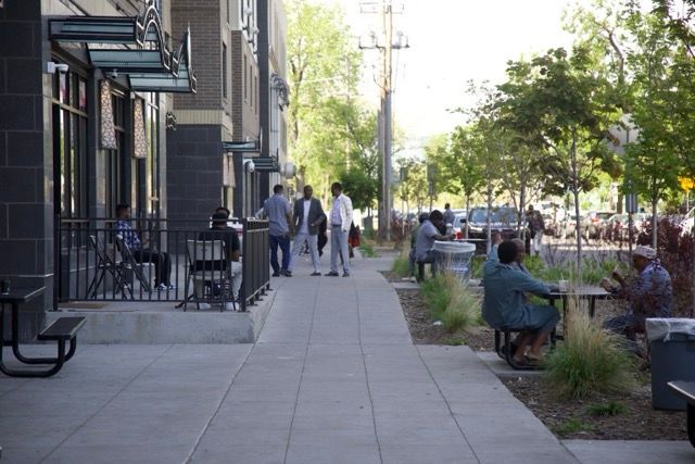 People outside a building with outdoor seating and trees on a sunny day.