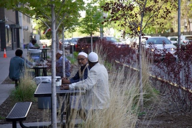 Men in traditional white clothing sit at outdoor tables, conversing near trees and parked cars.