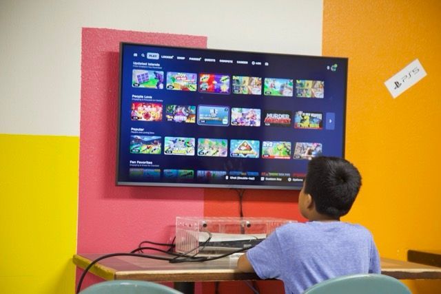 Boy seated at a desk looking at a large TV displaying a video game menu; colorful walls.