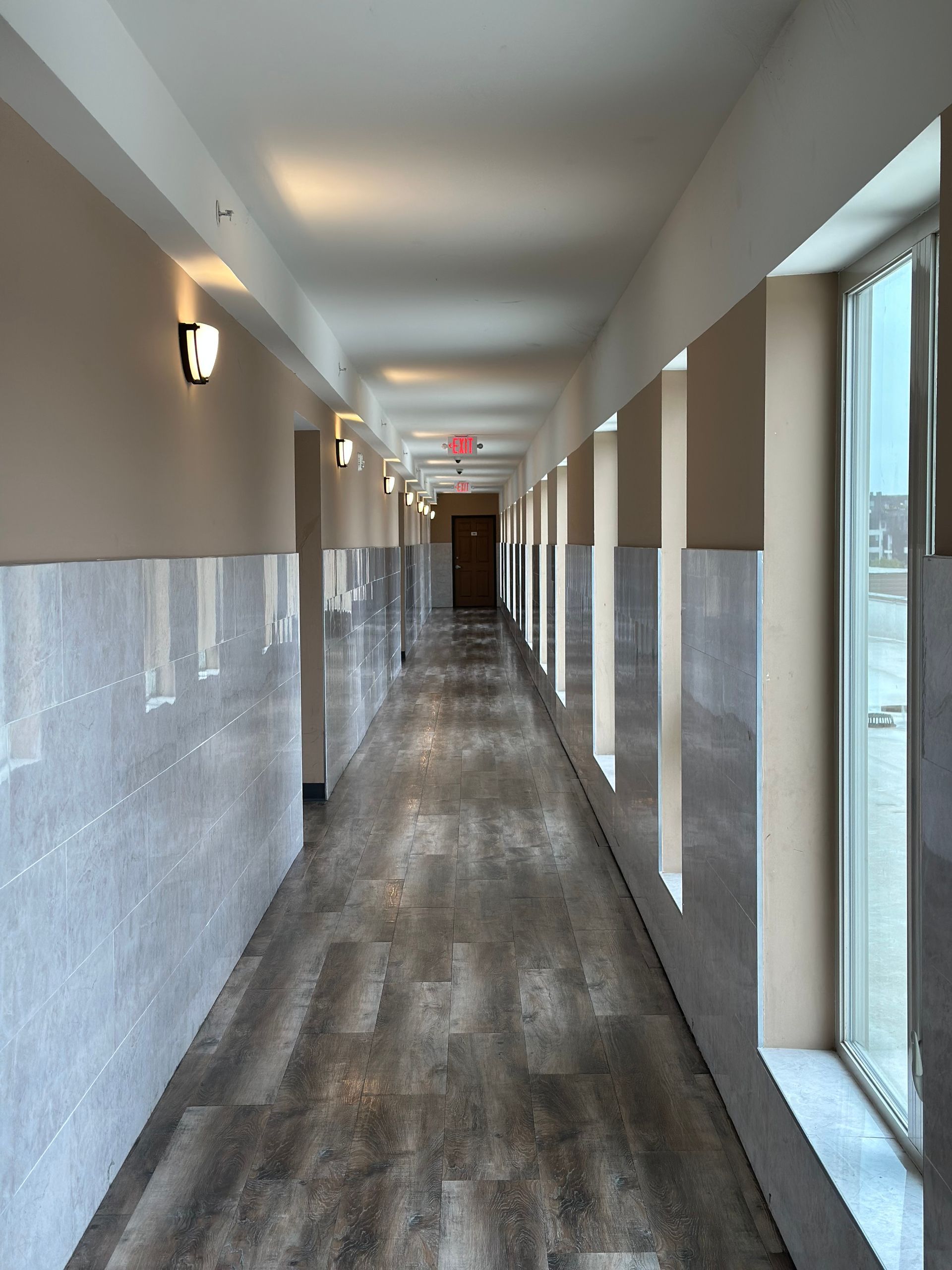Long hallway with wood-look flooring, windows, and recessed lights. Gray and beige walls, exit sign at the end.
