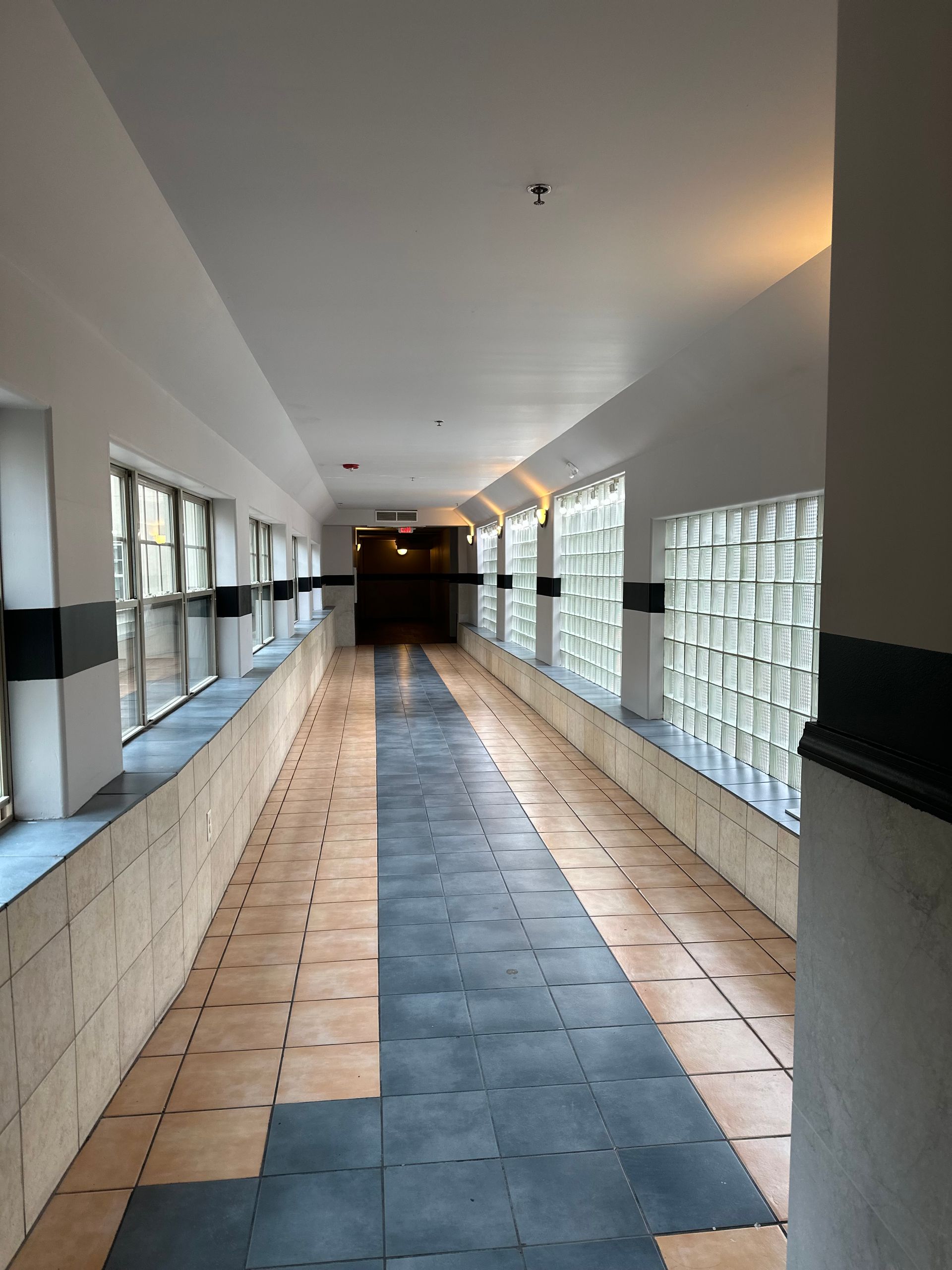 Long hallway with windows. Grey and tan tiled floor, windows with glass block, white walls, dark doorway at the end.