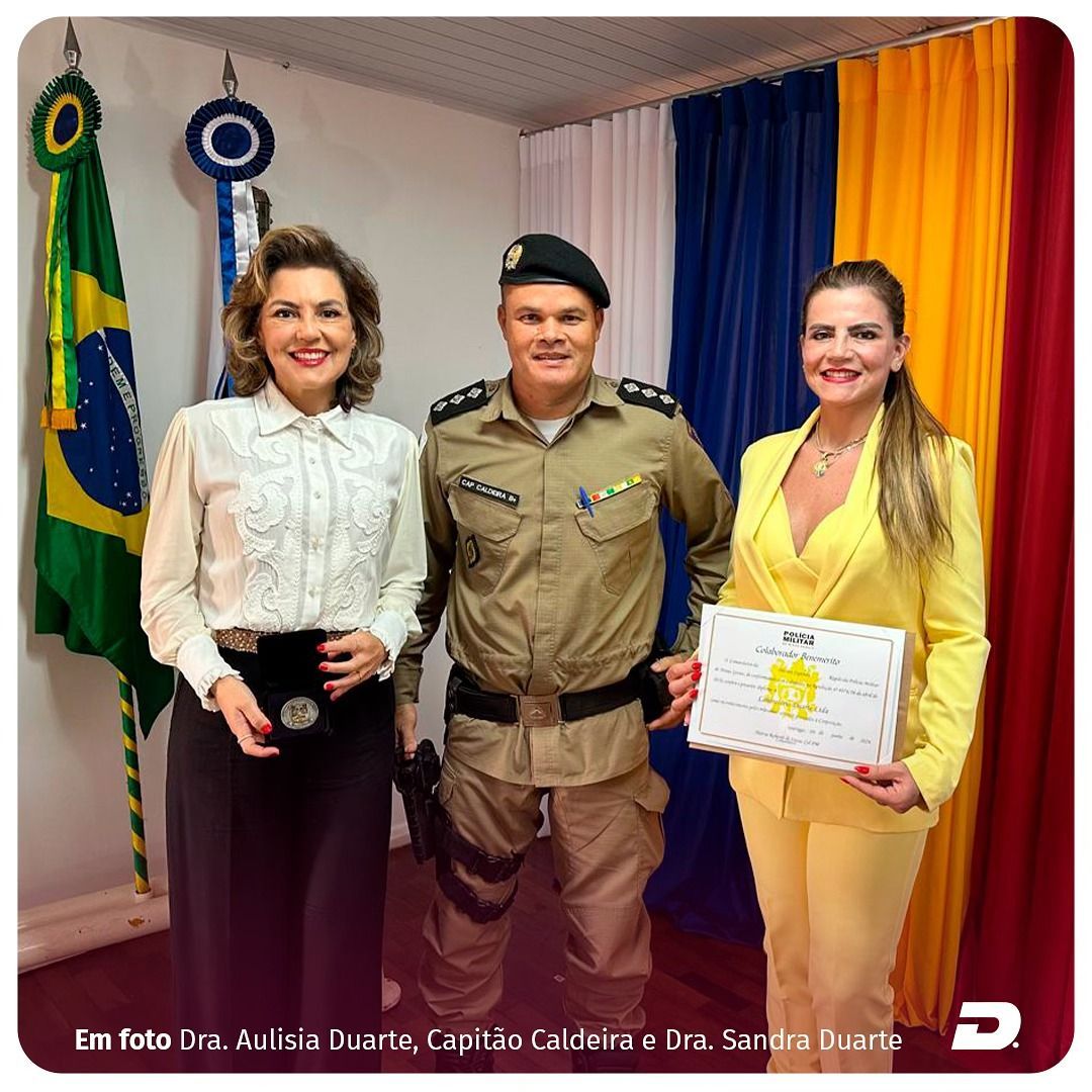 Três pessoas posam em um ambiente interno diante de bandeiras; um homem fardado está entre duas mulheres, uma delas segurando um certificado.