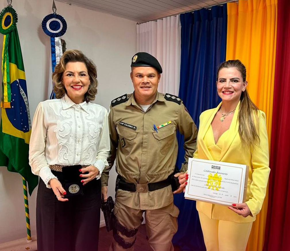 Três pessoas posando em uma cerimônia de premiação, segurando um certificado e em frente a cortinas coloridas.