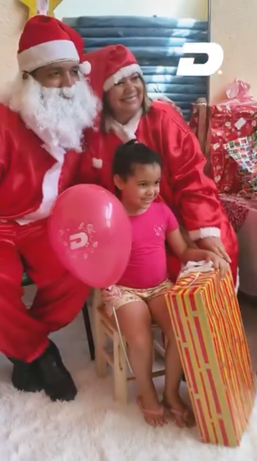 O Pai Natal e a Mãe Natal agacham-se ao lado de uma criança que segura balões cor-de-rosa e uma caixa de presente num ambiente festivo.