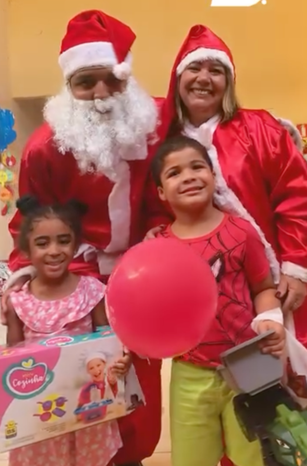 O Pai Natal e uma mulher posam com duas crianças que seguram presentes e um balão cor-de-rosa num evento festivo de Natal.