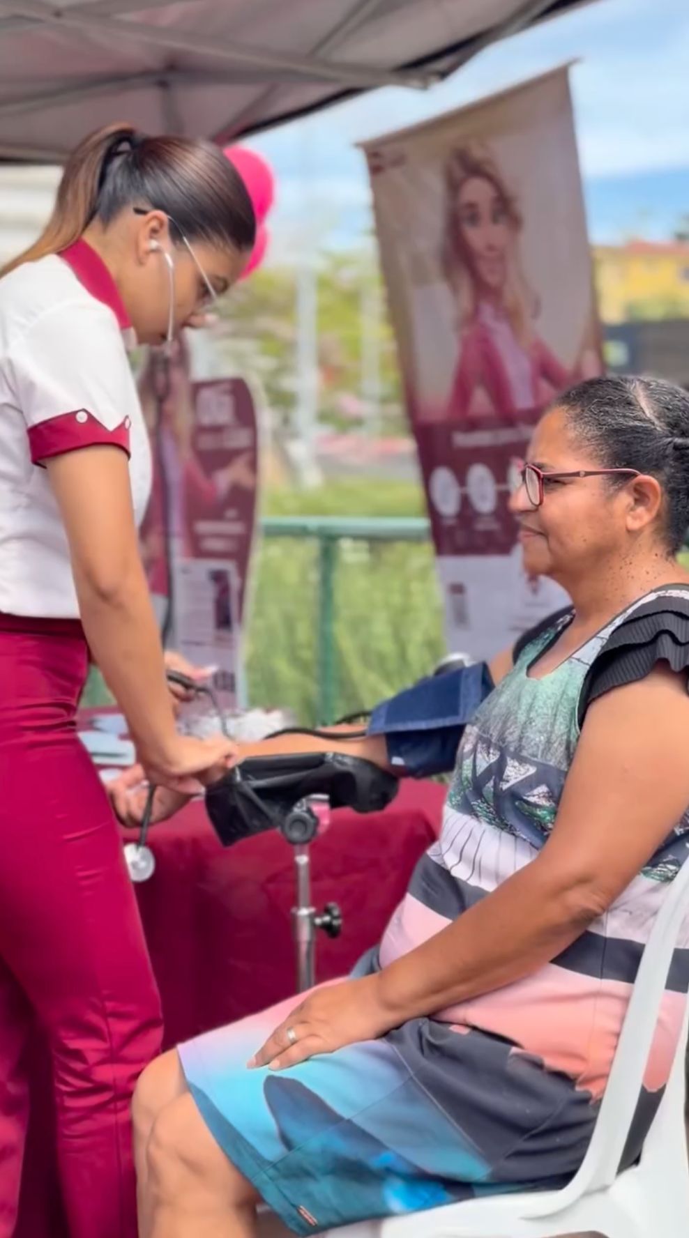 Duas mulheres conversando em um posto de saúde ao ar livre, uma sentada em uma cadeira branca e outra em pé, usando uniforme vermelho.