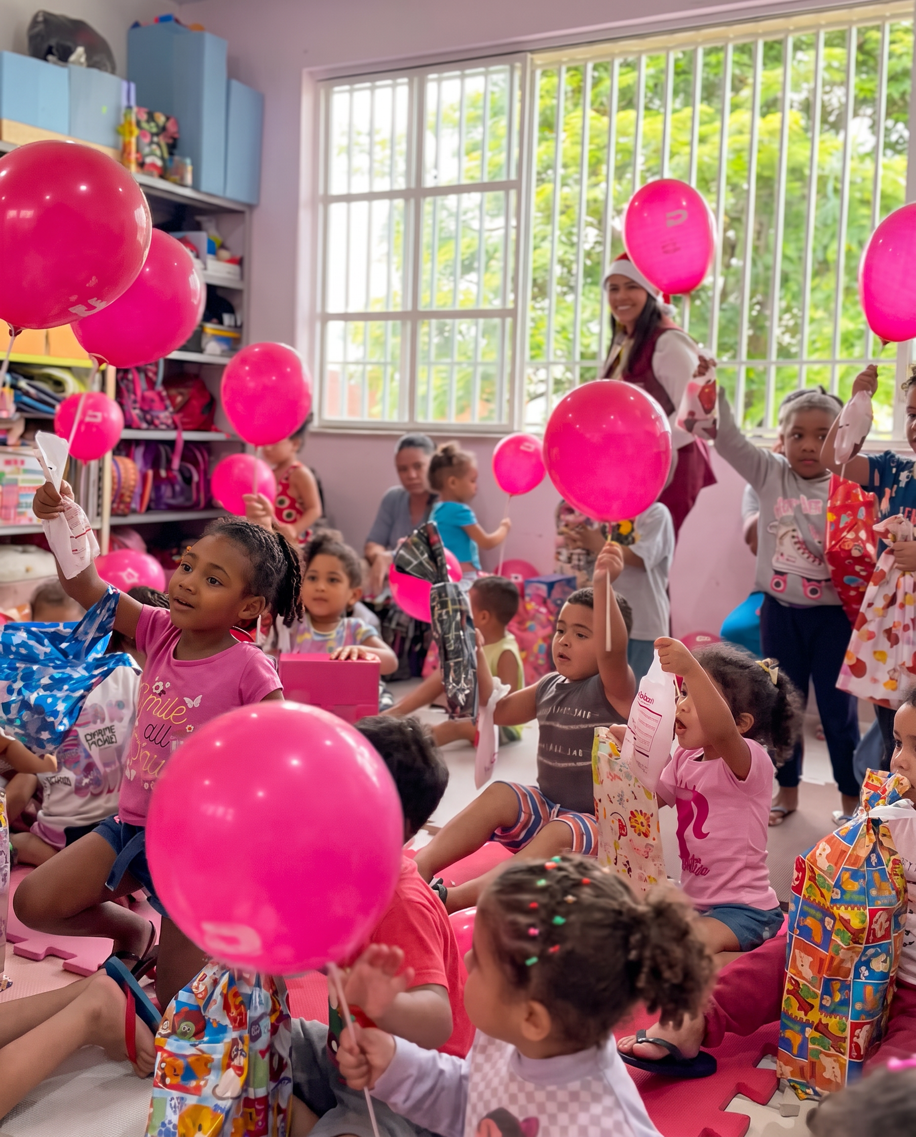 Crianças a celebrar com balões cor-de-rosa numa sala luminosa com grandes janelas