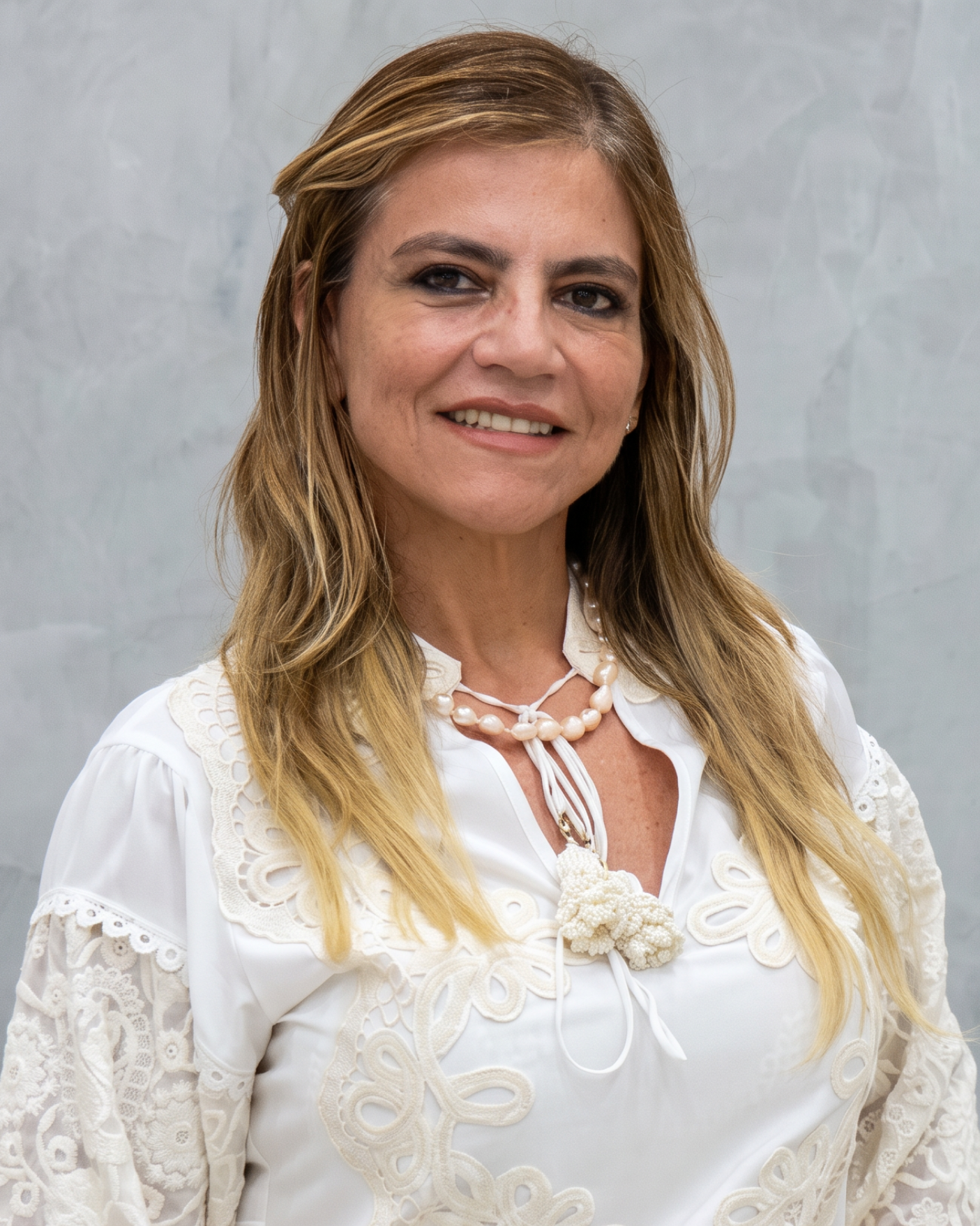 Mulher sorridente com uma blusa branca com detalhes em renda, em pé diante de um fundo cinza.