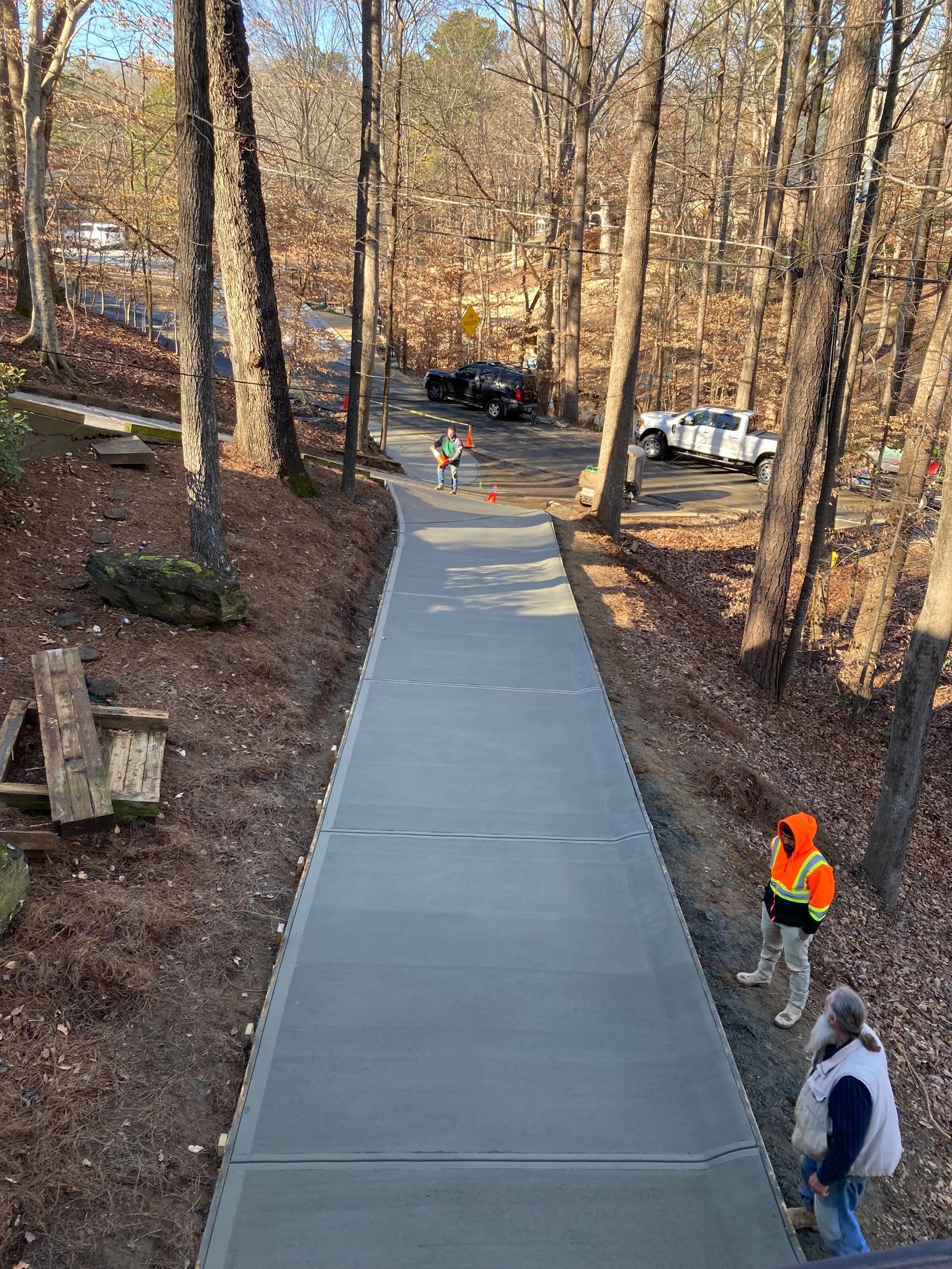 Newly poured concrete driveway with workers. Forest setting with trees, trucks, and orange safety vests.