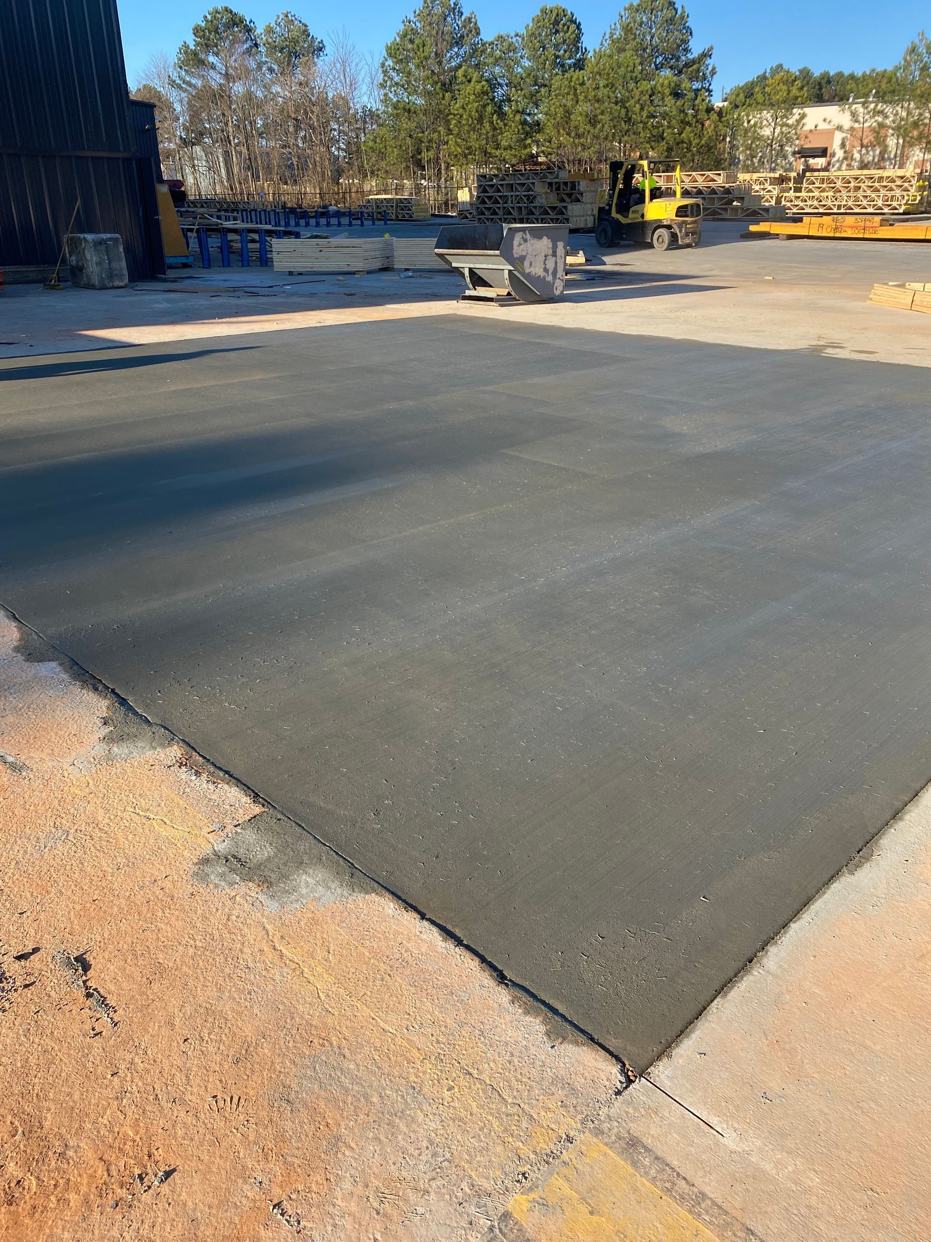Freshly paved asphalt area next to concrete. Lumber and equipment are in the background under a blue sky.