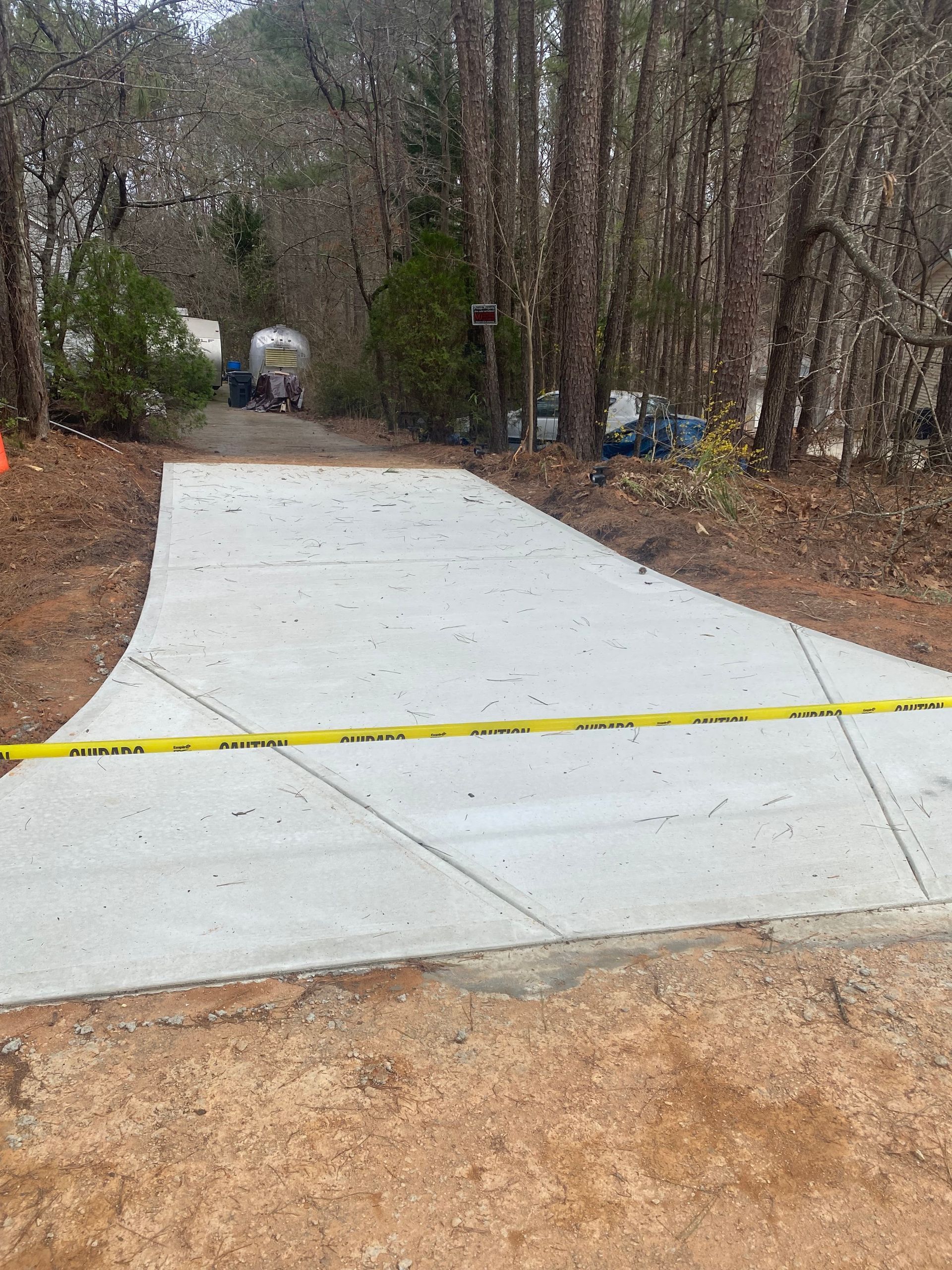 Newly poured concrete driveway, yellow caution tape across it. Trees and forest in the background.