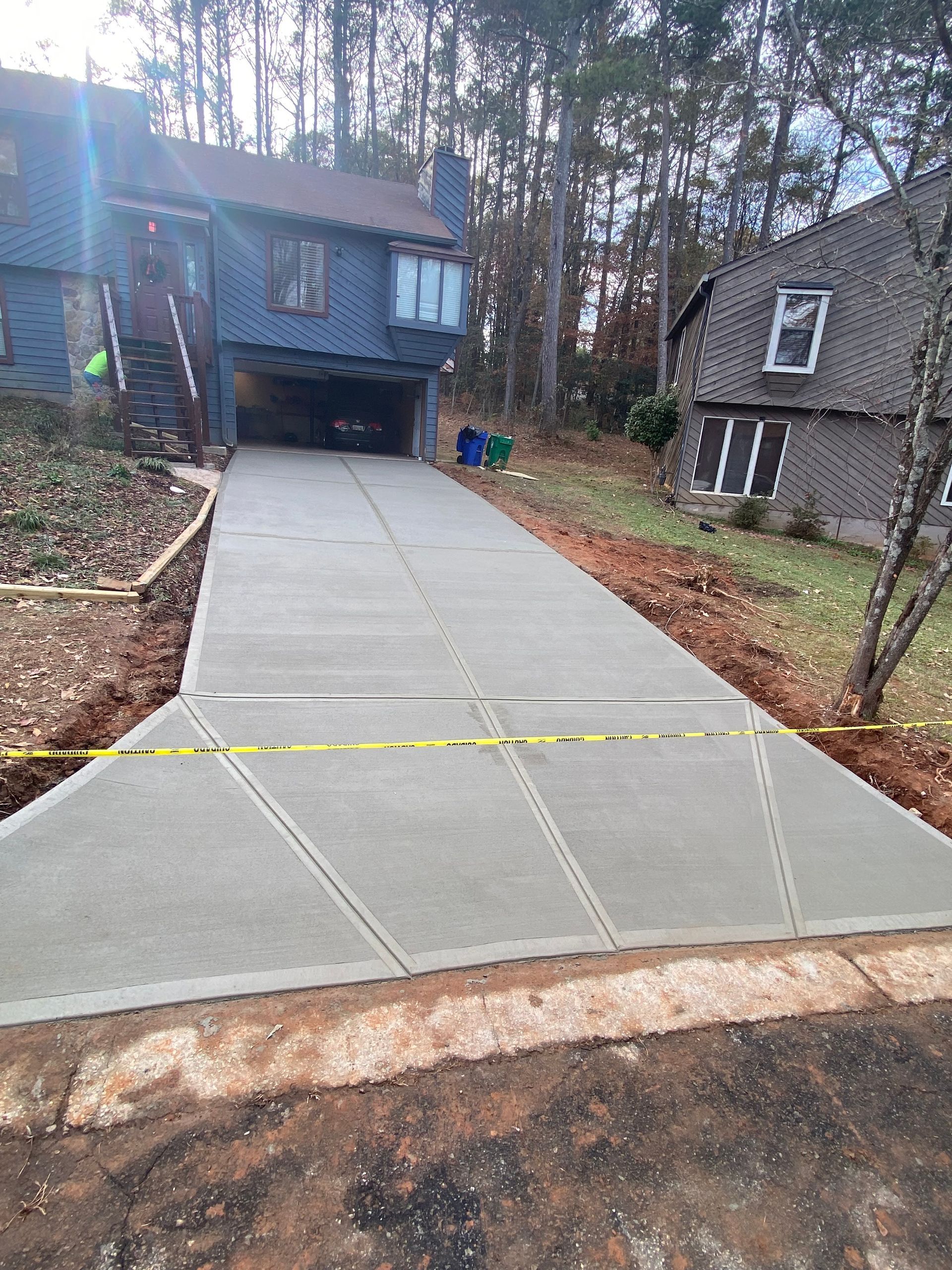 New concrete driveway in front of a blue house with attached garage; caution tape, trees in background.