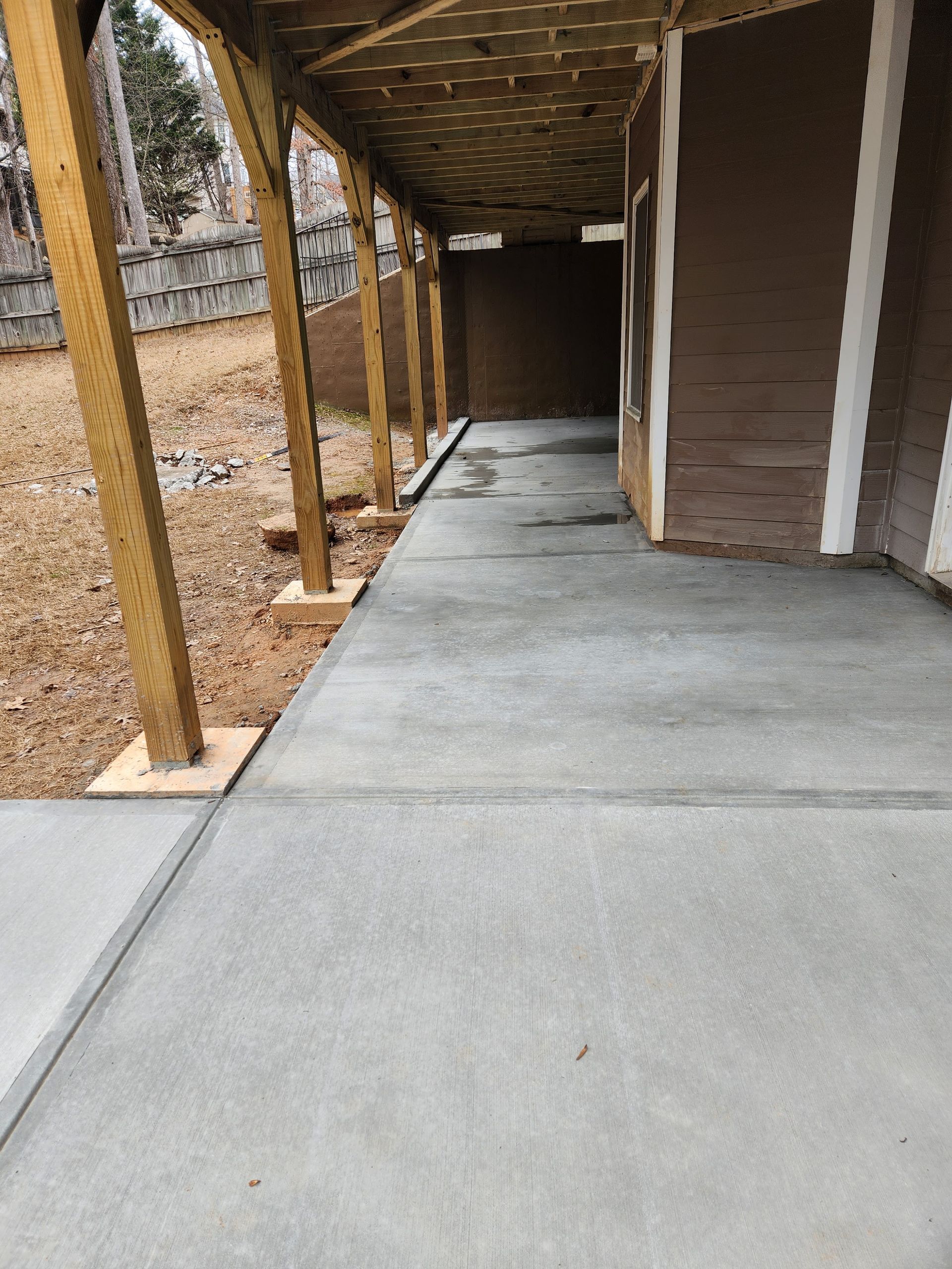 Concrete walkway under a covered porch with wooden support beams and a brown building.