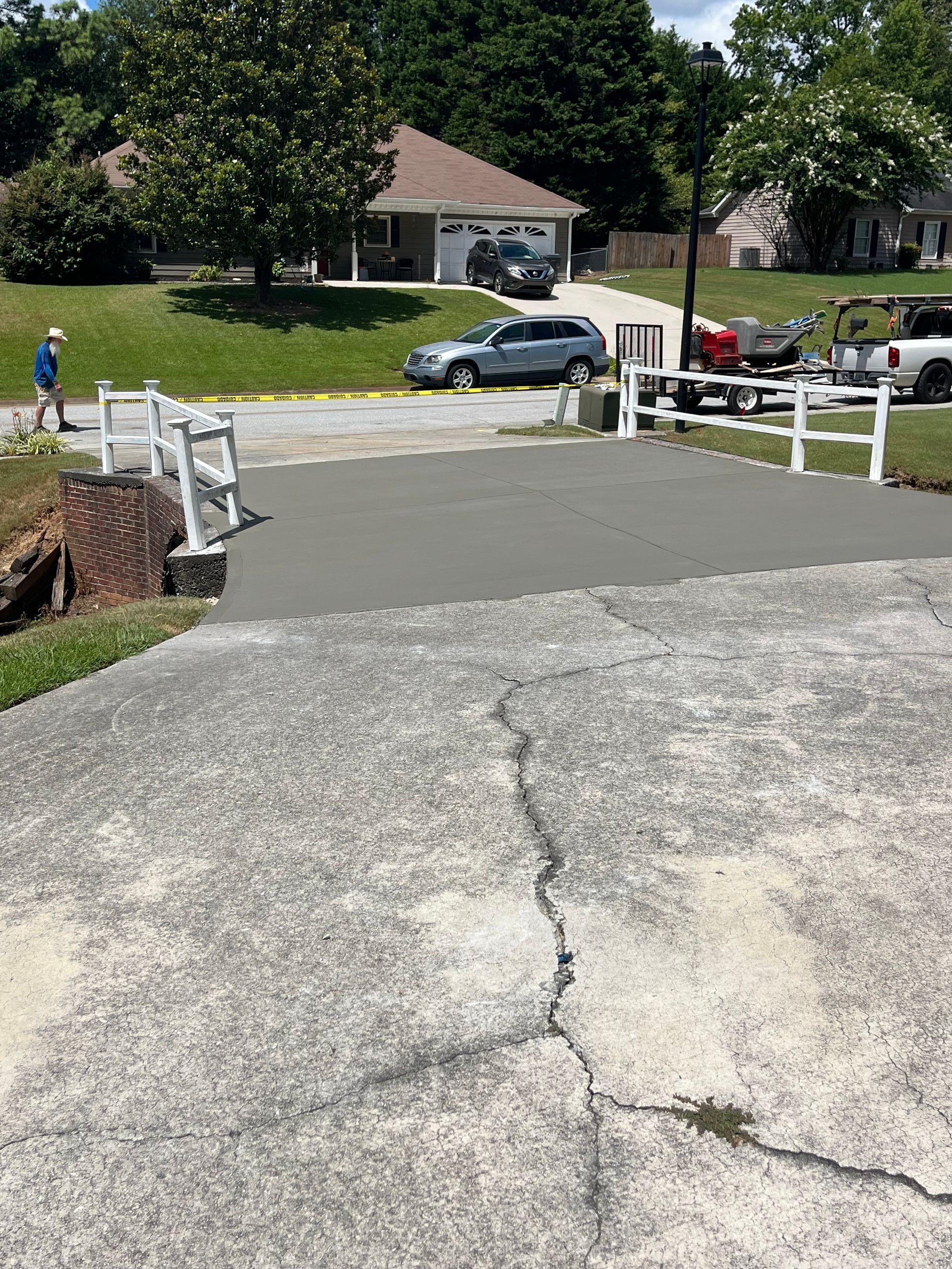 A freshly poured concrete driveway with white fencing. Old driveway with cracks. Cars and house in the background.