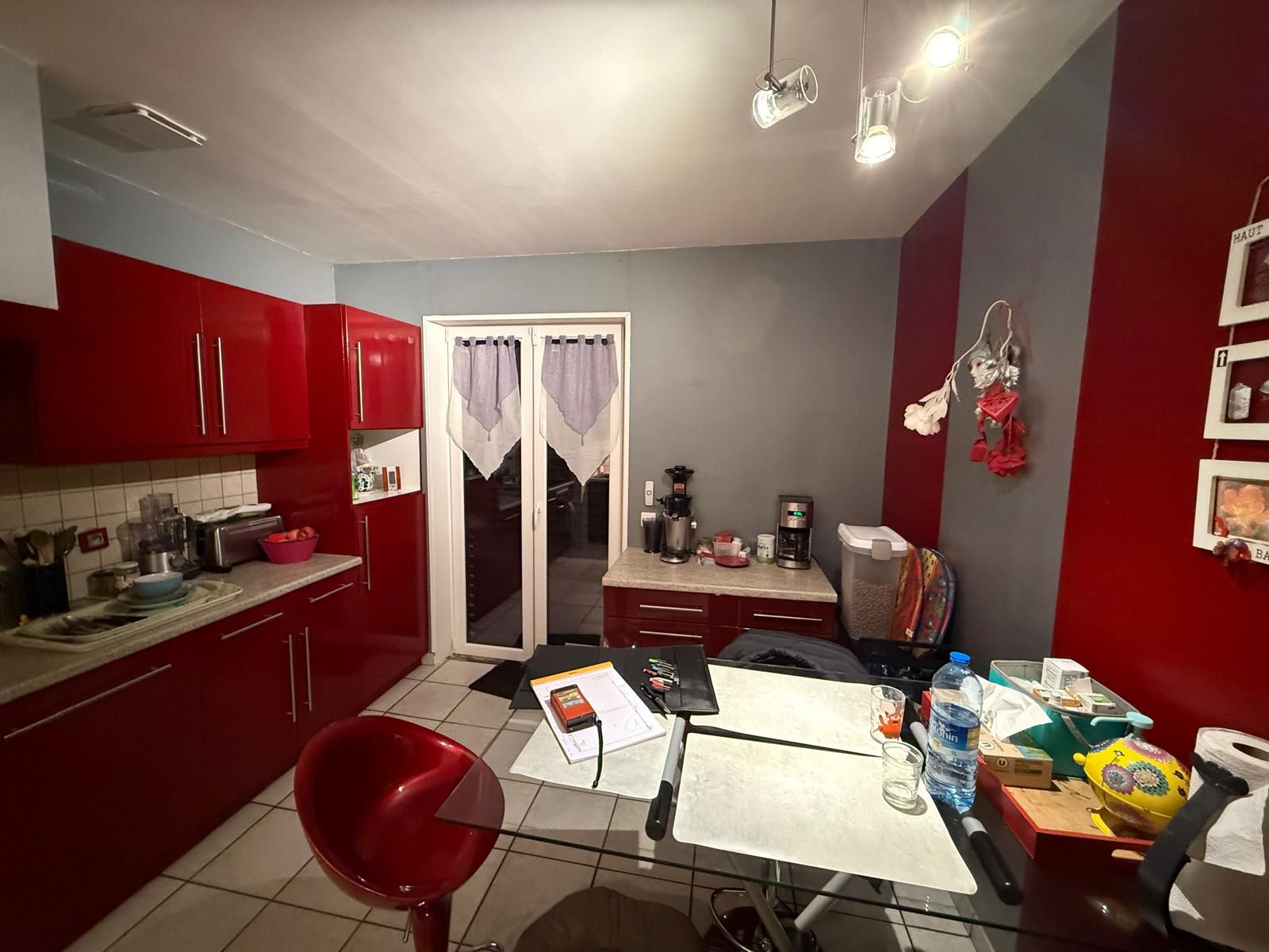 Cuisine moderne avec placards rouges, table blanche et portes vitrées donnant sur un patio sombre.