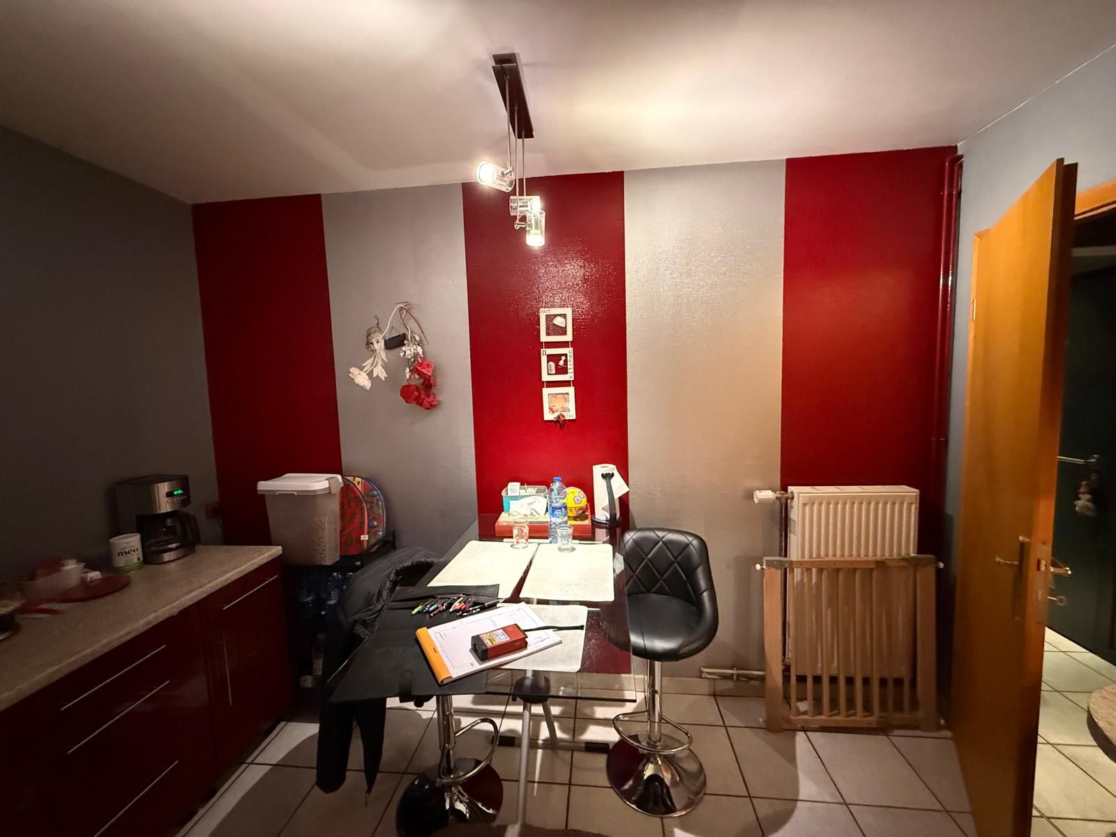 Petite pièce aux murs rayés rouges et blancs, avec un bureau, une chaise, des armoires et un radiateur près d'une porte ouverte.