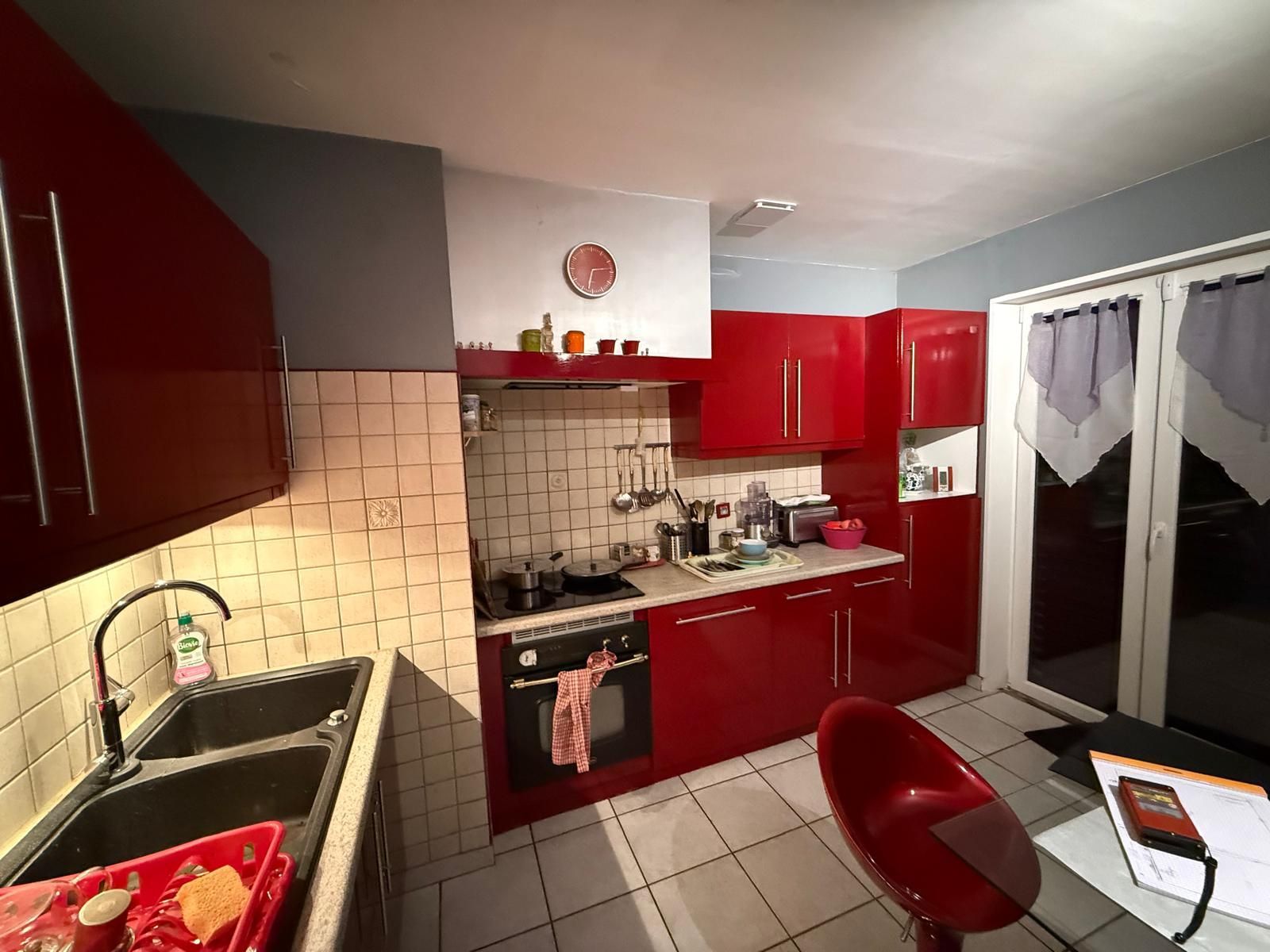 Cuisine rouge et blanche avec sol carrelé, évier, cuisinière, placards et portes vitrées s'ouvrant sur la droite.
