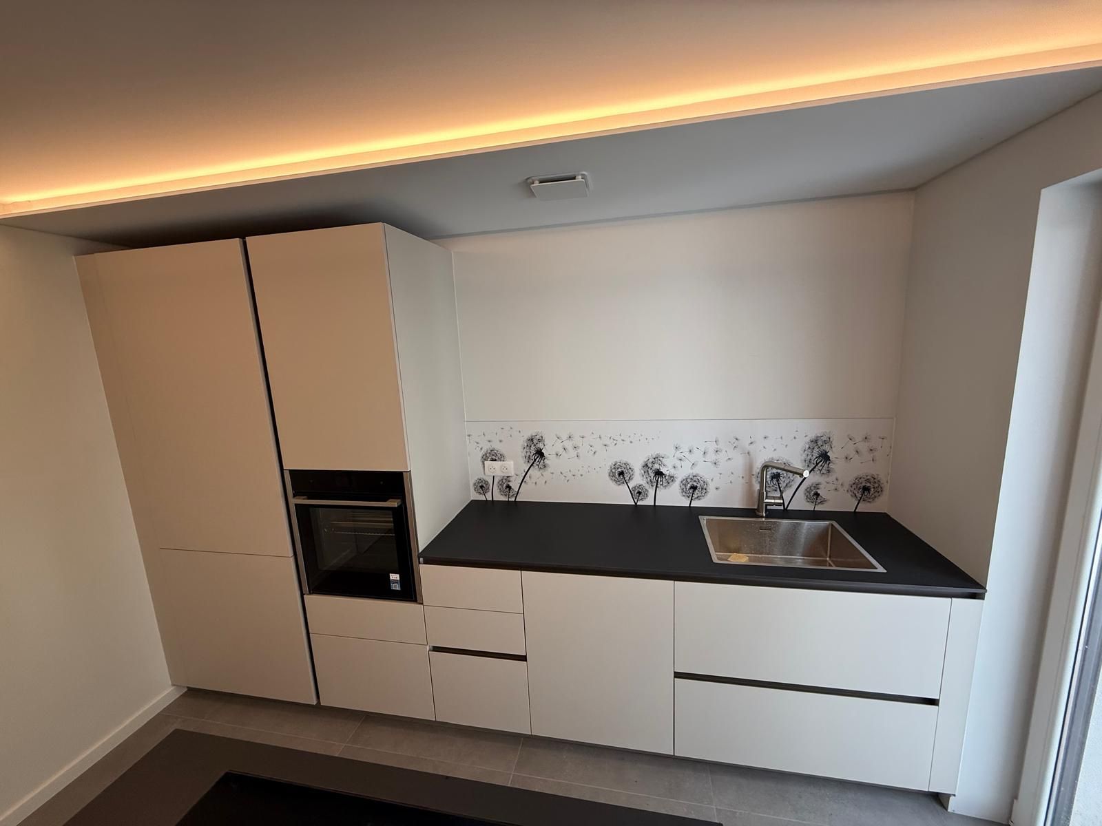 Kitchenette moderne et minimaliste avec placards beiges, plan de travail noir, évier et four encastré, sous un éclairage chaleureux au plafond.