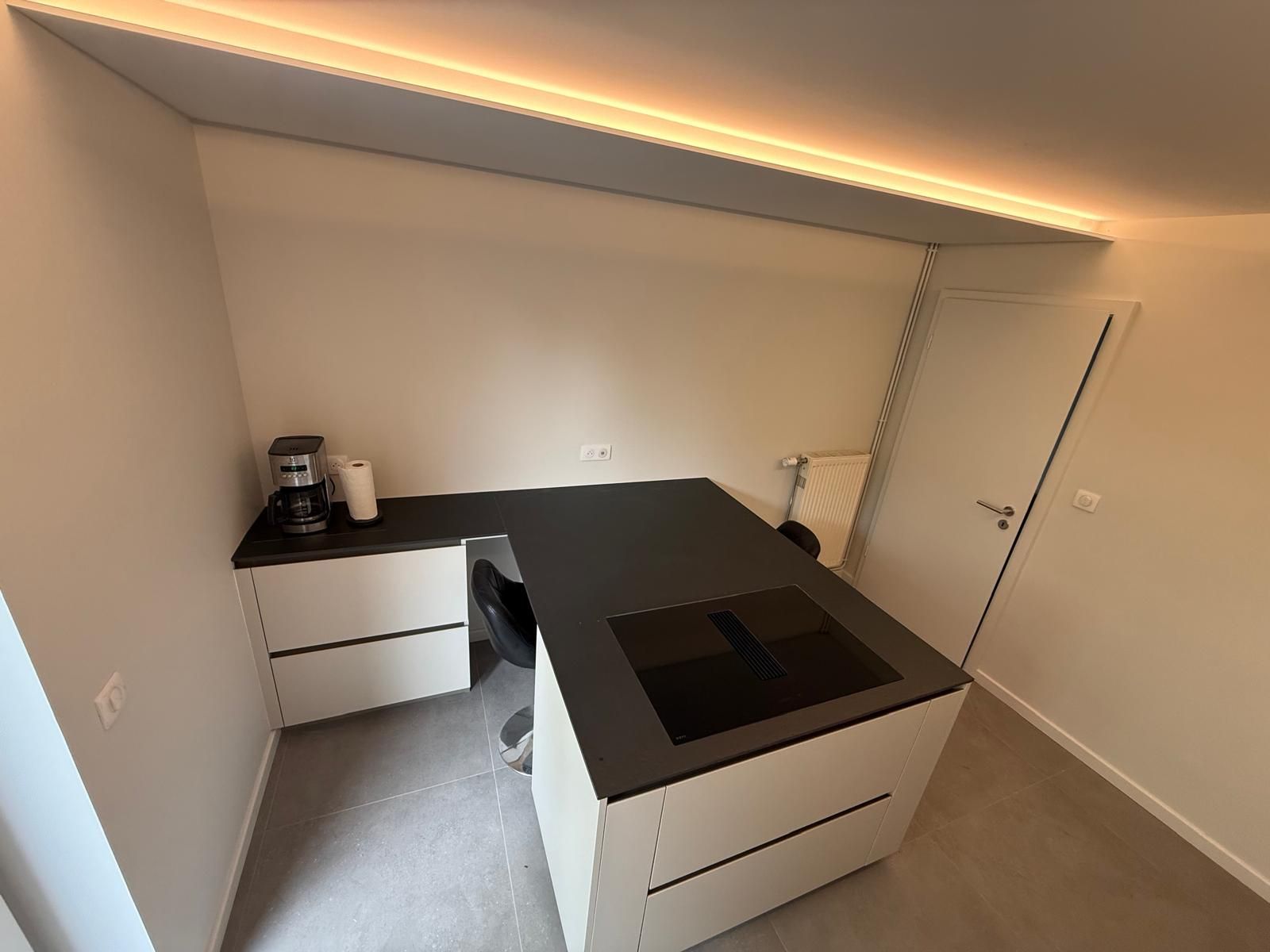Cuisine moderne compacte avec comptoirs noirs, armoires blanches et plaque de cuisson intégrée sous un éclairage chaleureux