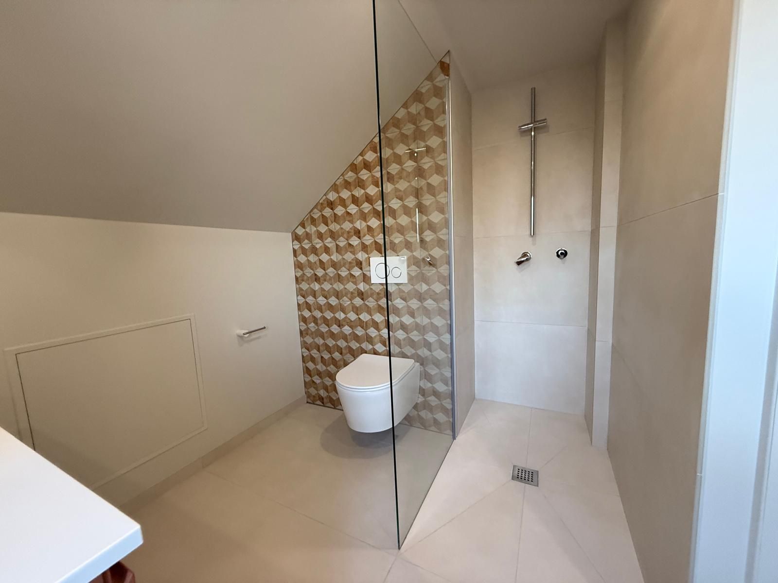 Salle de bains moderne avec paroi de douche en verre, toilettes blanches, sol carrelé et plafond mansardé