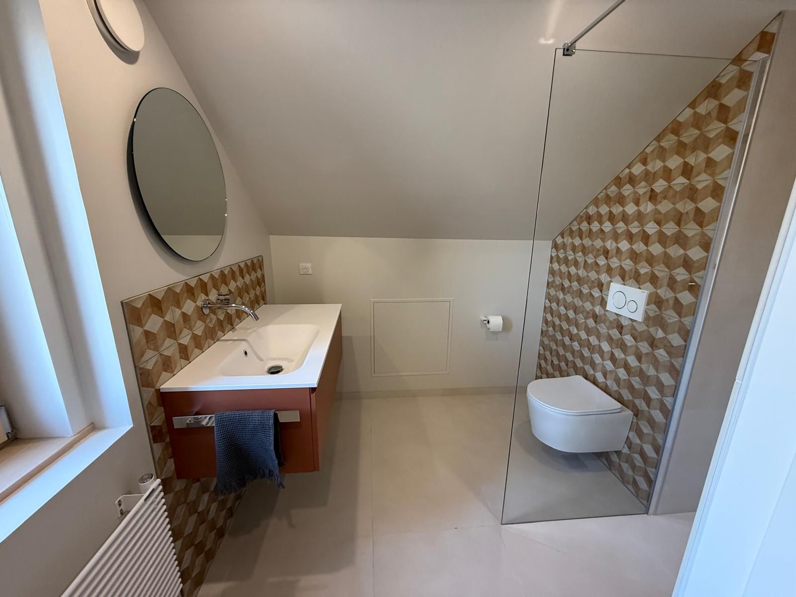 Salle de bains moderne avec lavabo mural, miroir rond, toilettes et mur d'accent en carrelage marron à motifs.