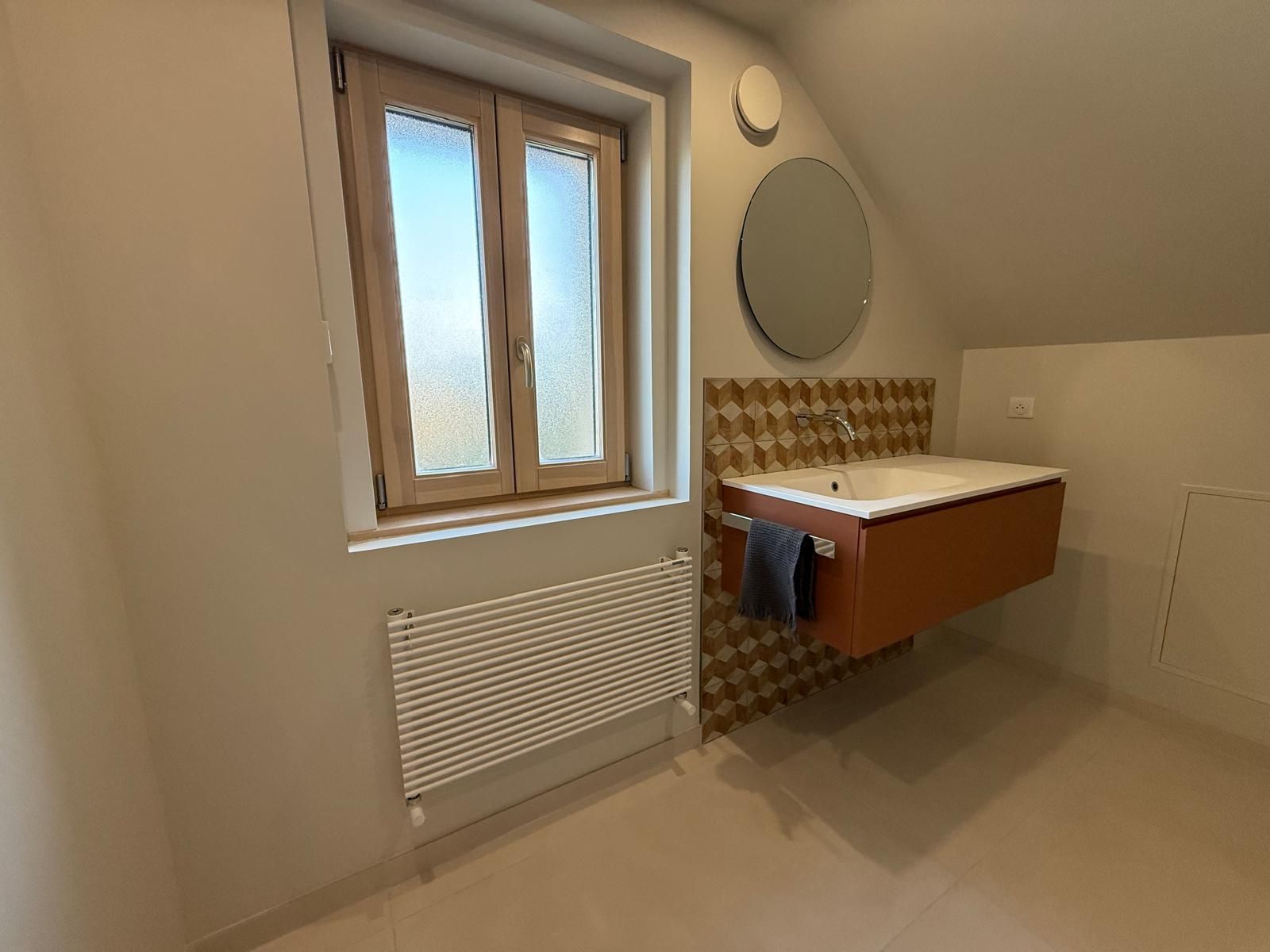 Petite salle de bains avec fenêtre, lavabo mural, miroir rond et radiateur sous la fenêtre.